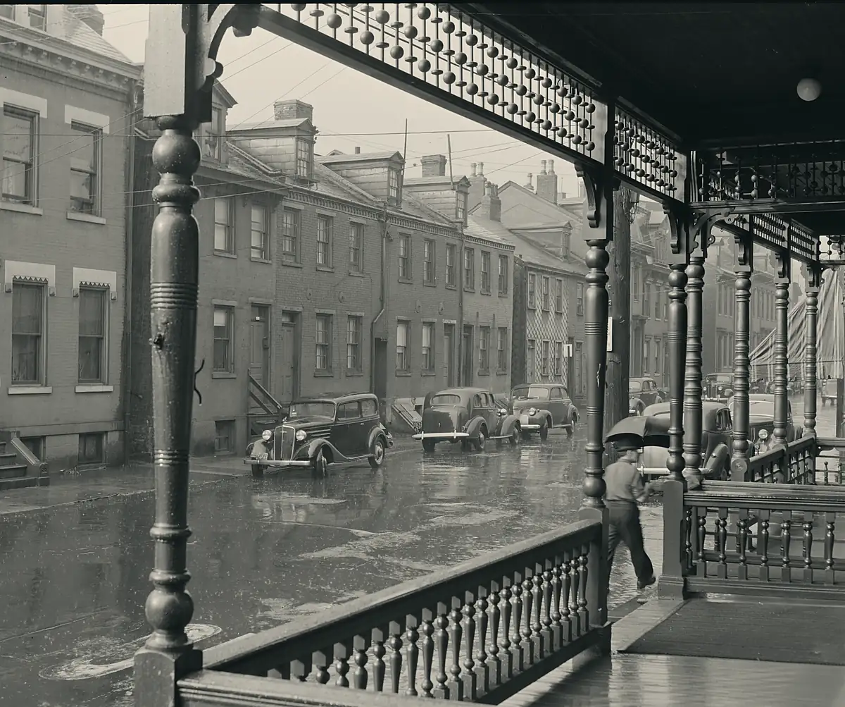Archival photograph: a wet street in heavy rain seen from a covered porch, water running off the roof edge above.
