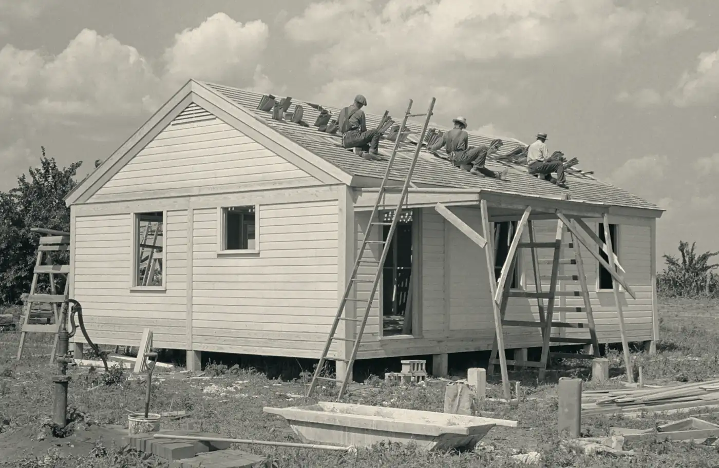 Archival photograph: three men laying shingles along the edge of a new house roof, a wooden ladder leaning against the eave.